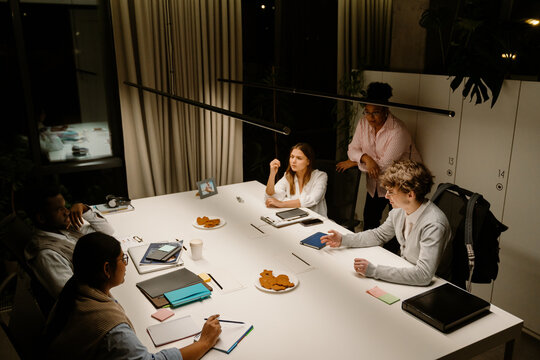 Multiracial group of young adults sitting and standing around the large white table during an evening meeting in a modern office. People discuss ideas, write notes, and use tablets and folders.