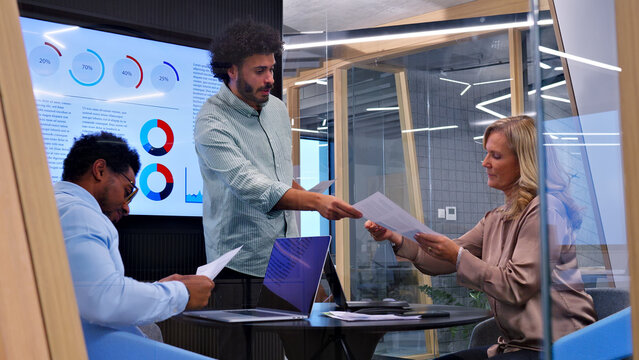 In a modern office setting, a man hands a document to a woman, symbolizing collaboration and teamwork. A seated man observes, highlighting communication and shared goals.
