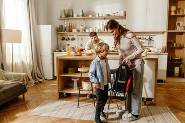 White adult man and woman helping a school-aged boy prepare for school in a bright kitchen, with the woman handing him a backpack while the man watches from the counter.