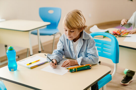 A school-aged White boy sits at a desk in a classroom drawing with colored pencils. The child is focused on his schoolwork during a creative activity in a modern learning environment.