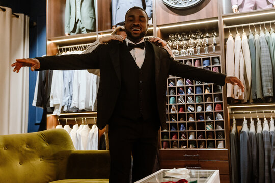 A Black man in his 30s is smiling with arms outstretched while being fitted for a tuxedo in a stylish suit store, standing in front of racks of jackets, shirts, and ties.