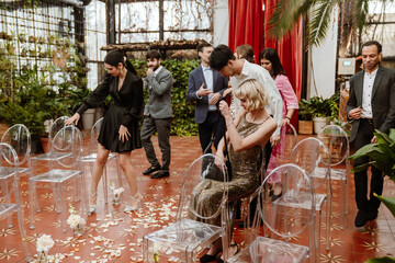 A multinational group of young wedding guests is walking and socializing in a decorated indoor wedding venue with floral arrangements and transparent chairs while in a lush, greenery filled setting.
