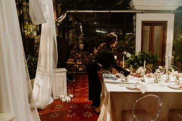 A White young female wedding planner with brown hair is setting up elegant wedding table in an indoor venue, arranging flowers, candles, and tableware in preparation for a beautiful celebration.