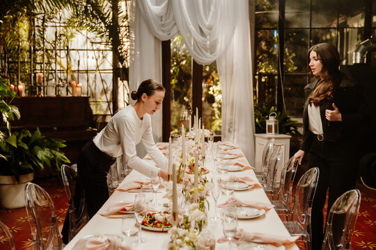 A White young waitress arranging a table for a wedding reception, while a White young female wedding planner with a black blazer observes the set-up in a decorated indoor venue with plants.