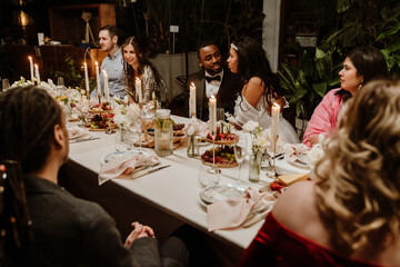 A Black man in a tuxedo and a woman in a white dress sit at the center of a decorated dinner table surrounded by guests during an elegant wedding celebration with candles, flowers, and food.