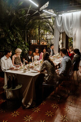 A Black groom and bride sitting at the head of a table with a multinational group of young wedding...