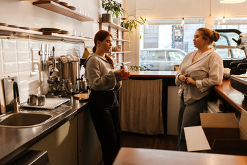 A White young female cafe owner and a White young female worker with red hair discussing renovation plans in a cozy cafe kitchen with modern equipment, preparing for the upcoming cafe reopening.