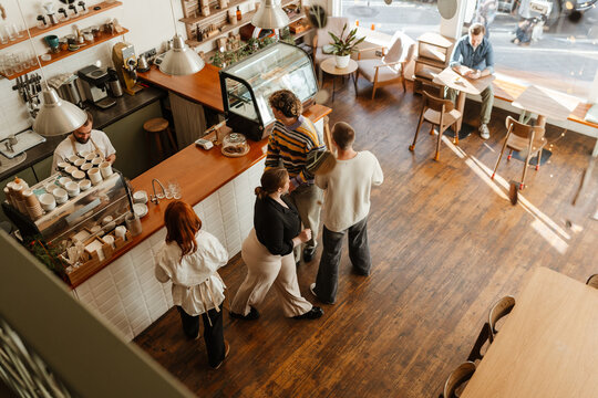 A group of adults queuing at the counter in a modern cafe while a barista prepares coffee, with one man seated by the window using a phone.