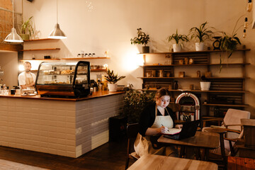 A White young female cafe owner and a White male barista working at renovated cafe, barista preparing drinks, owner using laptop, cozy atmosphere, plants, modern decor, and a glass display case.
