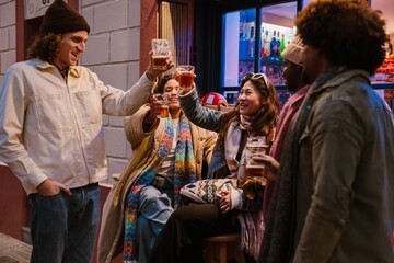 A multinational group of young people is laughing and enjoying drinks together at a bar, celebrating with beer glasses in hand, wearing cozy scarves and jackets, surrounded by warm lighting.