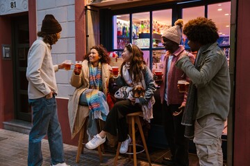 A multinational group of young friends enjoying drinks outdoors in front of a bar, laughing, having a great time together in the evening with a lively atmosphere and good company.