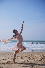 Elegant ballet dancer leaping on sandy beach in flowing pink dress under sky