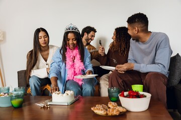 A multinational group of young people and men celebrating a birthday party at home, with a Black young woman wearing a tiara and pink feather boa, serving cake and enjoying the moment with friends.