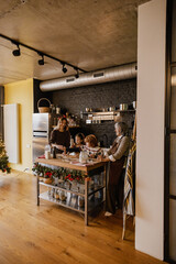 A White adult blonde woman, her mother, son, and daughter baking Christmas treats in a cozy modern kitchen, surrounded by holiday decorations, a Christmas tree, and festive kitchen tools.