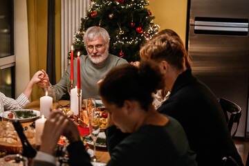 A White elder man and his family holding hands during a Christmas dinner, sharing a special holiday moment together around the festive table, with a decorated Christmas tree in the background.