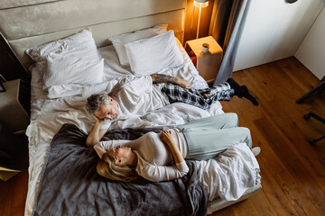 A White elder man with a tattoo and his wife with blonde hair, laying together in bed, enjoying a peaceful moment while smiling at each other in their cozy bedroom.