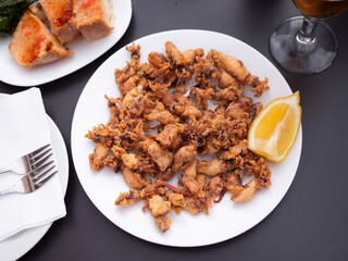 There is large portion of whole squid carcasses fried in batter in plate. Hearty seafood snack with beer and wine. Dish is made from deep fried squid clams and complemented with slice of juicy lemon