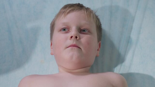 Sick young patient lying barechested on medical bed blinks multiple times, opens and closes mouth gently during examination, portraying subtle discomfort and quiet endurance in clinical setting