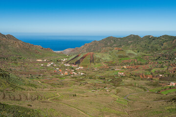 Obraz premium Panoramic view of a rural valley with terraced fields and scattered houses in the mountains of Tenerife, Canary Islands, with the Atlantic Ocean visible in the distance under a clear blue sky