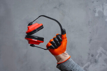 Naklejka premium A hand in an orange glove holds red protection headphones gear against a textured grey background. Concept construction safety and protection.