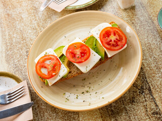 Fresh open-faced sandwich with tomato, avocado, and cheese on whole grain bread, garnished with aromatic dried herbs on rustic plate