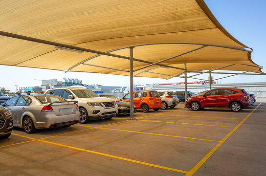 A row of vehicles is parked beneath large tan fabric shade sails that provide protection from the sun in an outdoor parking lot. The canopy structure features metal support posts and covers car park.