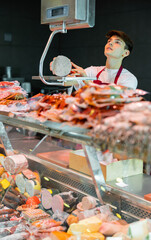 Young man in apron working in butchery, standing at counter, using scale to weigh sausage.