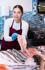 Portrait of friendly female fishmonger showing fresh fish to woman behind counter of seafood store