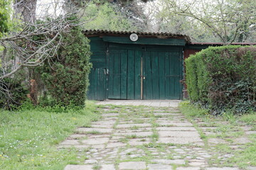 Old green wooden garage with weathered doors and overgrown path in Petrovaradin Fortress, Novi Sad, Serbia. Rustic storage building surrounded by spring vegetation and uncut grass.