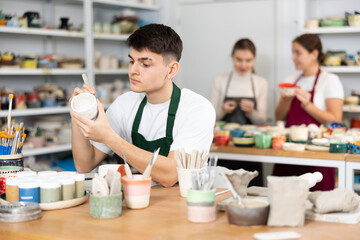 Concentrated young man in apron engraving on clay cup with cutter in pottery workshop