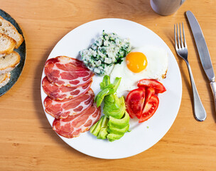 On table made of natural wood there is plate with healthy breakfast.On plate of fried eggs,risotto with spinach, slices of smoked bacon supplemented with chopped avocado and tomato