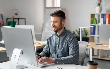 Cheerful middle aged businessman sitting at worktable at office, typing on computer keyboard,...