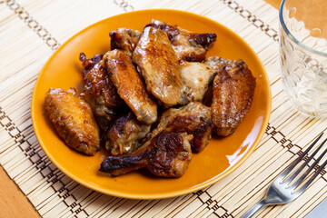 Hearty food - juicy and fatty fried chicken wings. On set table there is plate with fried bird wings, cutlery, wine glasses, spices