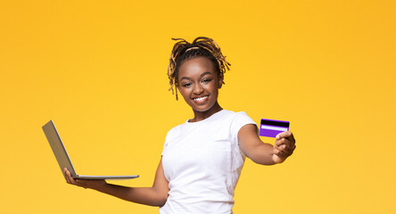 Easy online shopping. Happy african woman with laptop showing credit card, yellow studio...