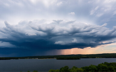 Naklejka premium Dramatic storm cloud formation fills the sky, looming over a green treeline and dark water. A rain shower is visible under the imposing cloud mass in a natural outdoor setting.