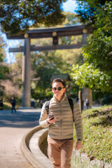 Fototapeta premium Latin young man exploring Meiji Shrine in Tokyo, wearing beige jacket, brown pants, sunglasses, and carrying a backpack with a cheerful attitude.