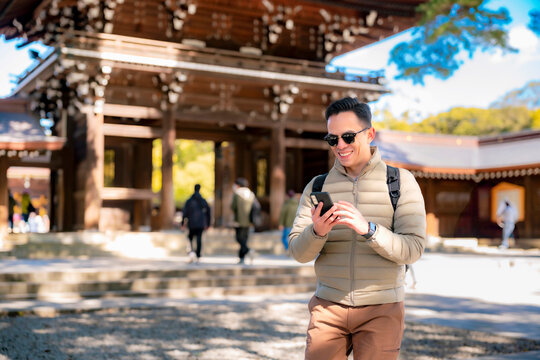 Young Latin man in beige winter clothes exploring Meiji Shrine in Tokyo, smiling with sunglasses, phone in hand, and relaxed attitude.