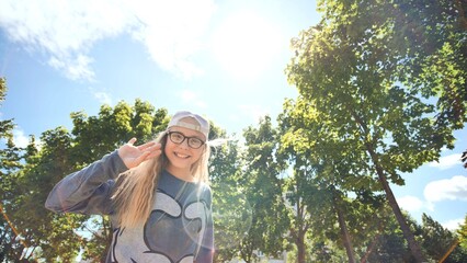 Cheerful schoolgirl wearing glasses, cap, waving happily in sunlit park during summer afternoon