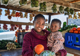 Happy African street vendor holding her child, shack with vegetables and fruits, women empowerment