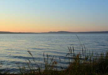 View through the reeds to the calm surface of a large lake surrounded by hills just after sunset.