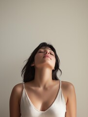Fototapeta premium Portrait of a young woman with her eyes closed and her head tilted upwards. she is wearing a white tank top with thin straps and her hair is styled in loose waves.