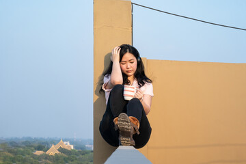 Glasses Asian woman is listening music via headset, drinking coffee on rooftop barricade cement...