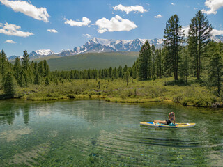 A man does yoga on a SUP board in a transparent lake in the forest, against the background of mountains, the tops of which are covered with snow. Mountain transparent lake in the Altai forest in autum