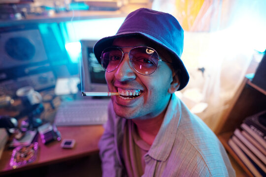 Close-up portrait of young male programmer with cigarette in mouth, wearing purple bucket hat with sunglasses, smiling for camera