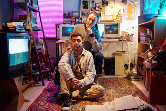 Young programmers posing in messy room with vintage computer monitors and CRT television, girl putting hand on mans shoulder while he sitting on floor