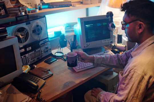 Young male programmer drinking coffee while writing code on retro computer, sitting at wooden desk with telephone, vintage tape recorder, books, empty cans and paper container on it