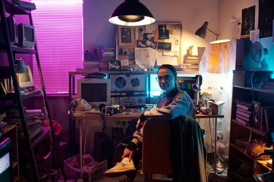 Portrait of Caucasian young woman posing for camera while sitting on chair in dimly lit messy room with vintage tech equipment