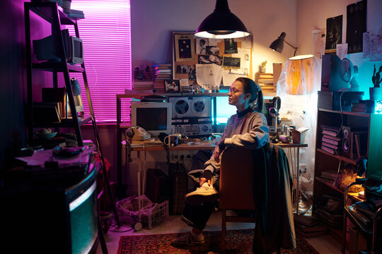 Portrait of Caucasian young girl posing for camera while sitting on chair in dimly lit messy room with vintage tech equipment