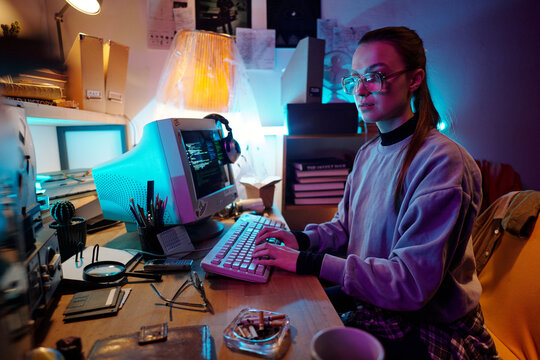 Caucasian young woman using old computer while sitting on chair in dimly lit messy room with vintage tech equipment