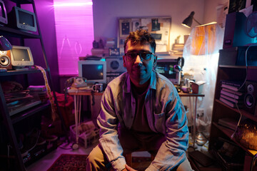 Portrait of young male programmer wearing glasses, shirt and pants stretching his neck and posing for camera in dimly lit room with retro devices and notice board in background
